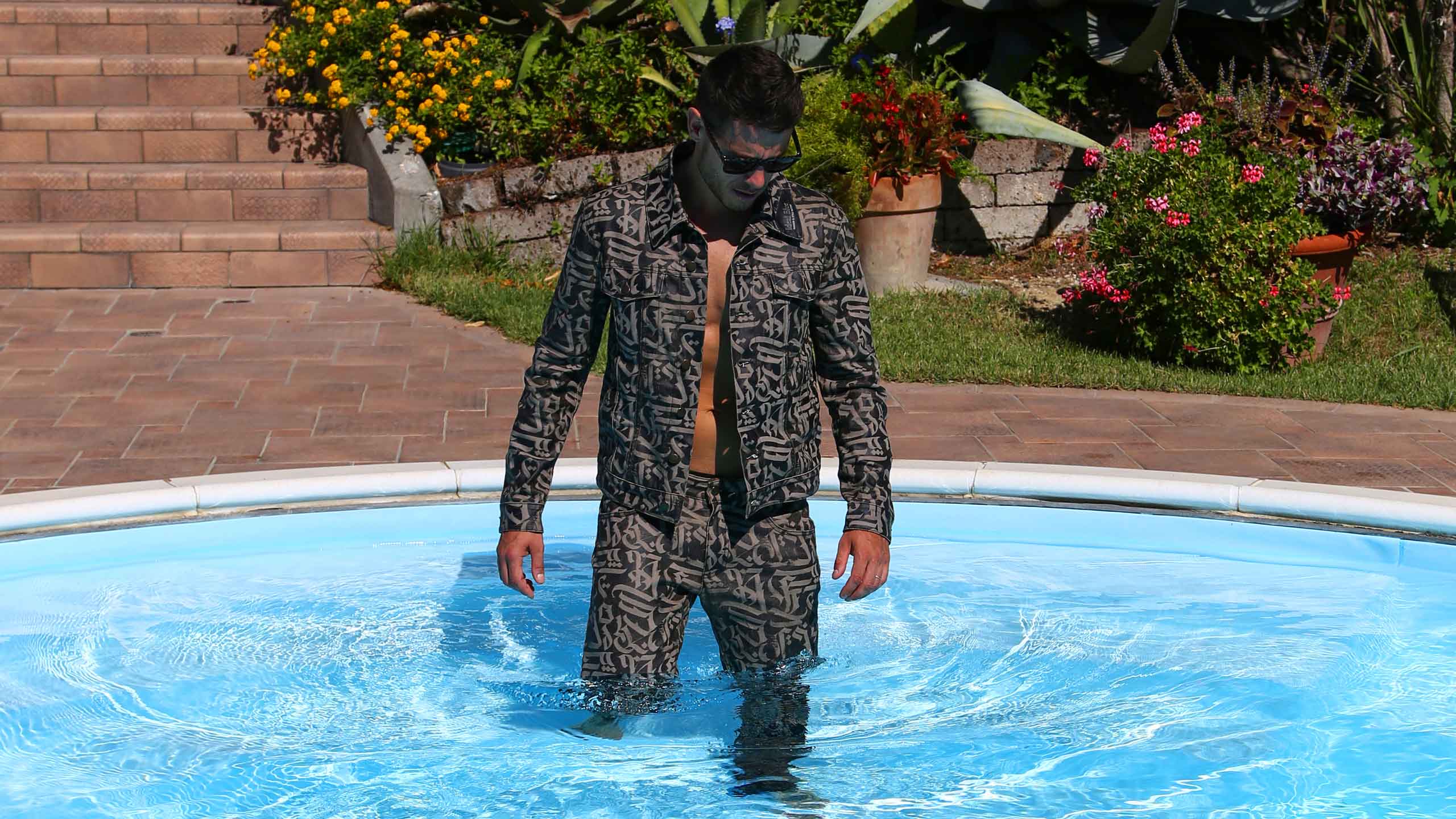 Shot of a Givenchy denim total look, customized with abstract calligraphy. The model is immersed in the pool and moving toward the room, looking at it.