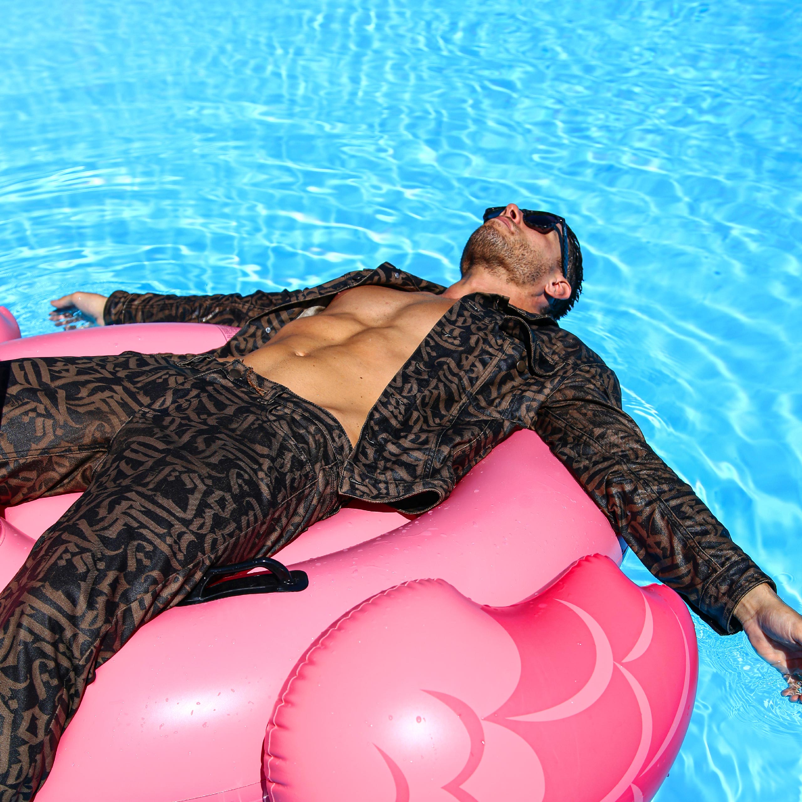Shot of a Givenchy denim total look, customized with abstract calligraphy. The model is in the pool, lying on a flamingo-shaped inflatable, with her arms submerged in the water.