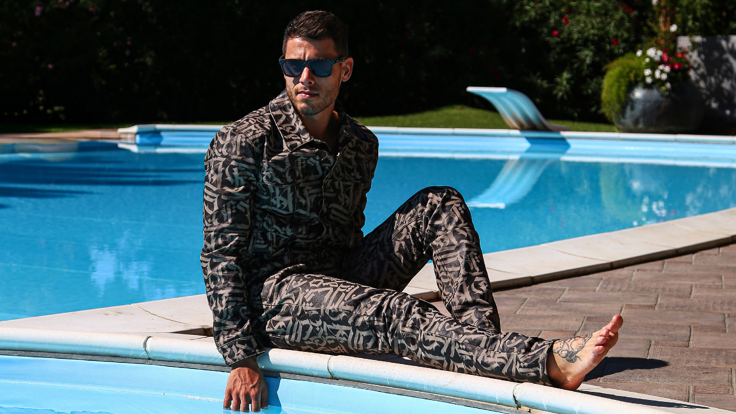 Shot of a Givenchy denim total look, customized with abstract calligraphy. The model poses poolside in a crouched position, with his right leg in the foreground.