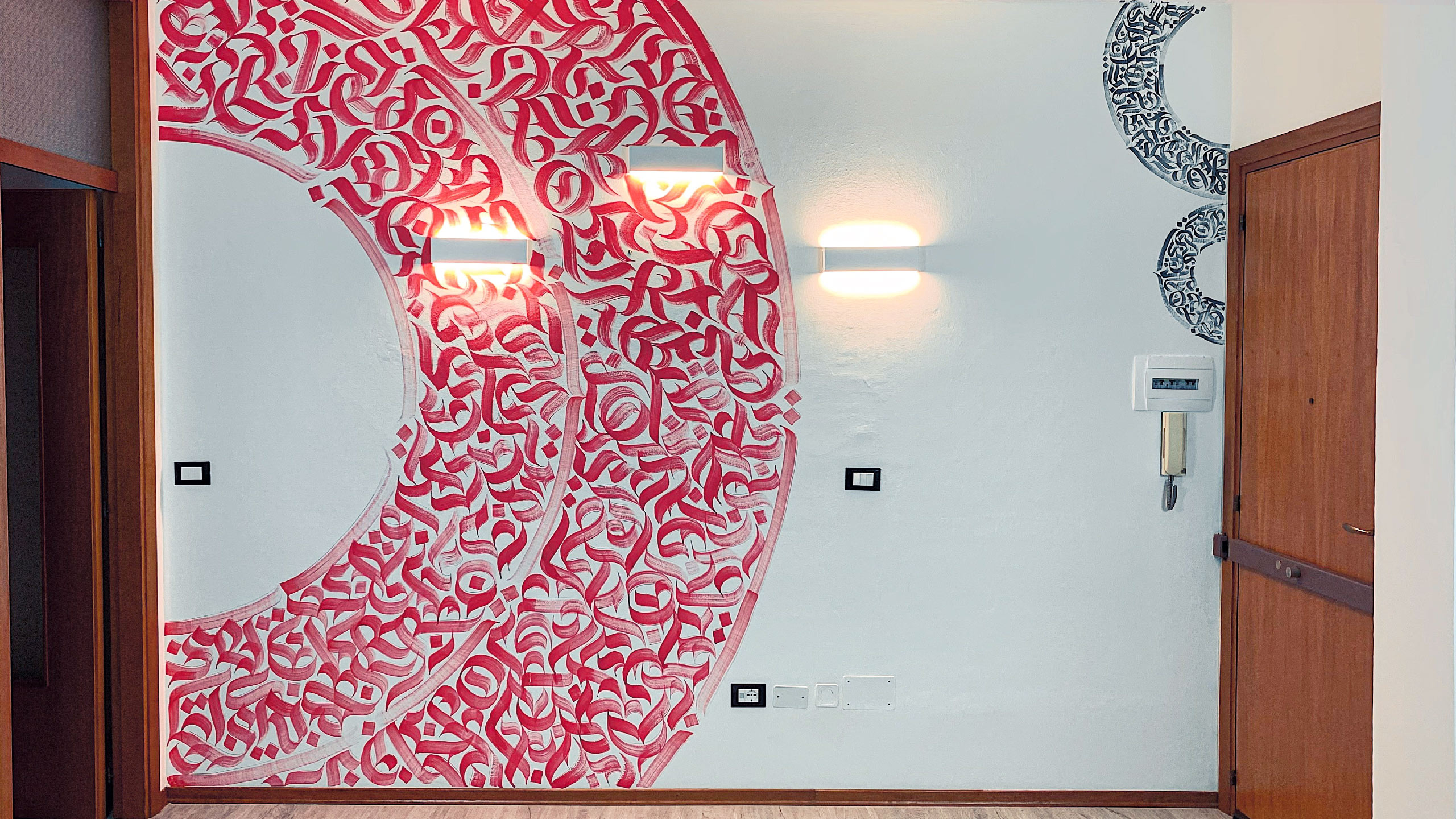 Front view shot of a part of the mural, at the corner of the living room of the private space, with the lights on and no frames on the walls. The mural represents a circumference composed of abstract calligraphy.